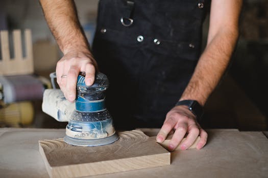 Unrecognizable male carpenter using random orbital sander while working with wooden detail at workbench in professional workshop on blurred background